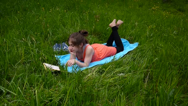 Una joven relajada leyendo el libro en el campo. Linda adolescente acostada en la hierba. Video filmación de alta definición de la cámara estática . — Vídeos de Stock