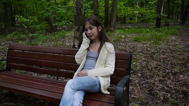 魅力的な少女は、ベンチで携帯電話に話します。夏の公園。笑顔。静的なカメラを撮影 Hd 映像. — ストック動画
