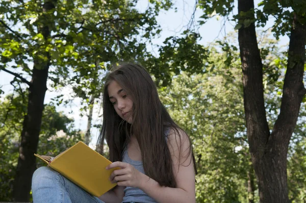 Linda adolescente leyendo libro sentado en el banco en el parque, estudiando al aire libre . — Foto de Stock