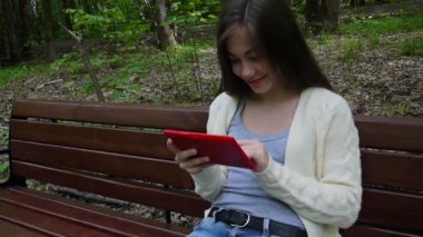 Güzel genç kız tablet bilgisayar ile parkta bankta oturur. Statik kamera film.