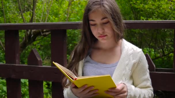 Retrato de adolescente sério lendo livro e virando página inclinado sentado no banco na floresta na primavera, estudando ao ar livre. Filmagem de vídeo em HD de câmera estática . — Vídeo de Stock