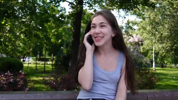 Ung vacker flicka talar till telefon på bänk. Sommaren park. Leende. HD video antal fot skytte statisk kamera. — Stockvideo