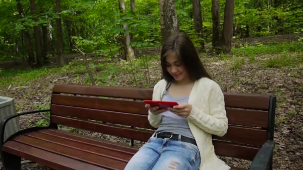 Güzel genç kız tablet bilgisayar ile parkta bankta oturur. Statik kamera film. — Stok video