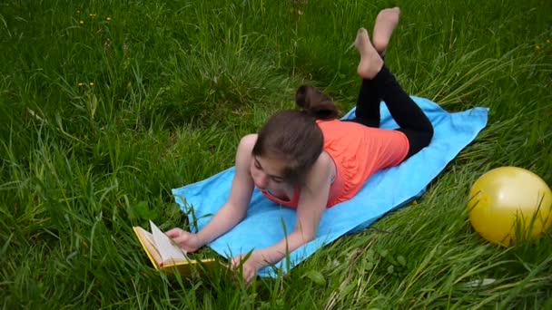 Ontspannen jong meisje het lezen van het boek in het veld. Leuke tiener liggend op het gras. Videobeelden Hd opnames van statische camera. — Stockvideo