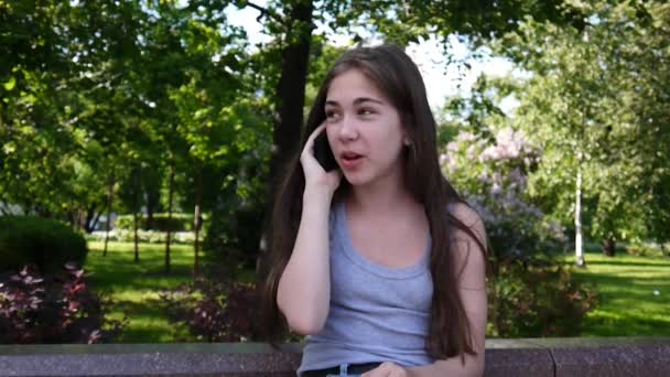 Молодая привлекательная девушка разговаривает по телефону на скамейке. Летний парк. Улыбнись Видеосъемка в высоком разрешении . — стоковое видео