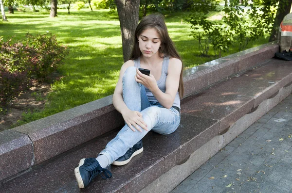 Молодая привлекательная девушка ждет звонка, сидя на скамейке. Летний парк. Фото — стоковое фото