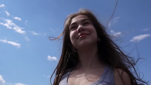 Close up portrait of smiling beautiful young girl on the blue sky background with blowing hair in wind — Stock Video