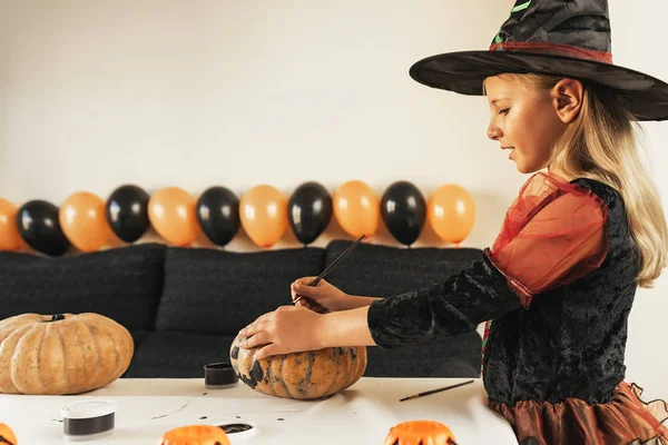 Hermosa chica disfrazada de bruja decorando una calabaza en casa . —  Fotos de Stock