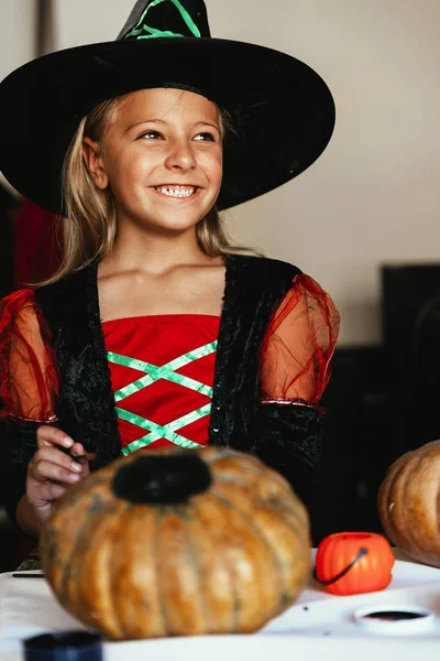 Hermosa chica disfrazada de bruja decorando una calabaza en casa . — Foto de Stock