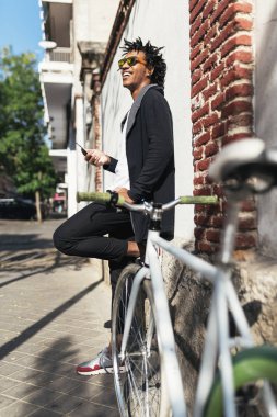 Cep telefonu ve sabit vites bisiklet kullanan Afro genç adam.
