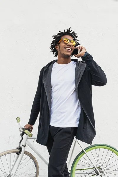 ชายหนุ่มแอฟโรใช้โทรศัพท์มือถือและจักรยานเกียร์คงที่ . — ภาพถ่ายสต็อก