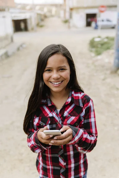 Portret van leuk latino meisje met behulp van de mobiele telefoon in sloppenwijk. — Stockfoto