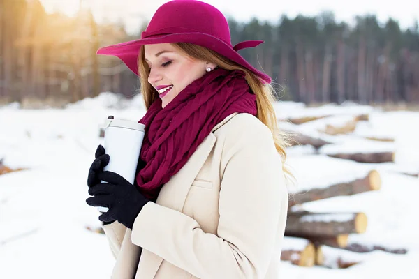 Bella, giovane ed elegante ragazza sorridente e mantenendo una tazza termo nella foresta. Moda . — Foto Stock