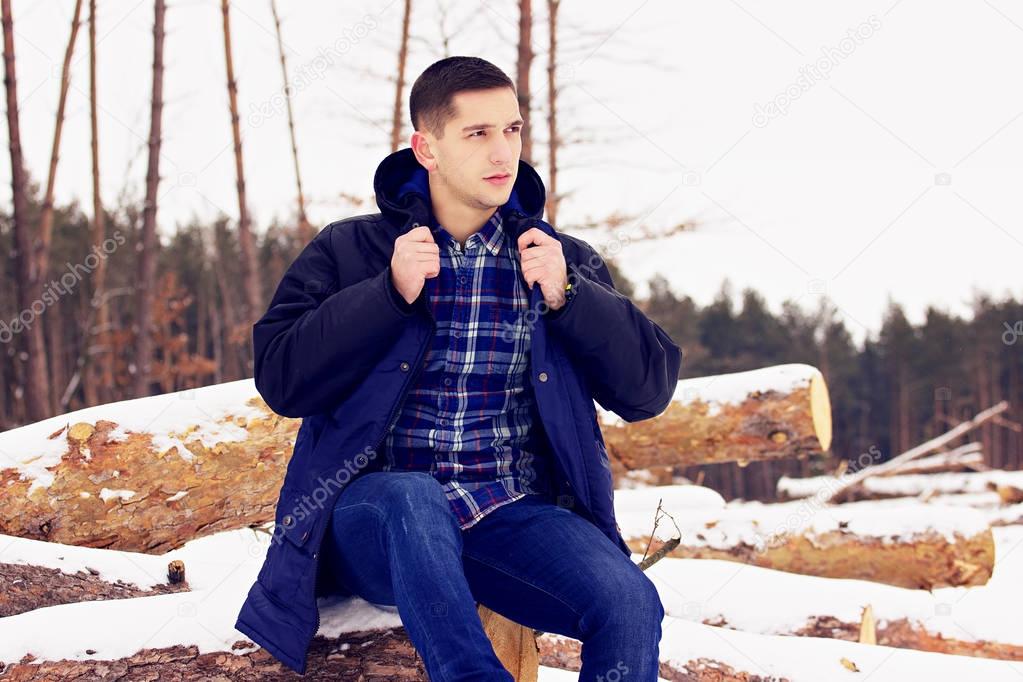 Handsome, stylish and young man correcting his jacket and sitting on the woods in the forest. Early spring, sunny day. Men's fashion.