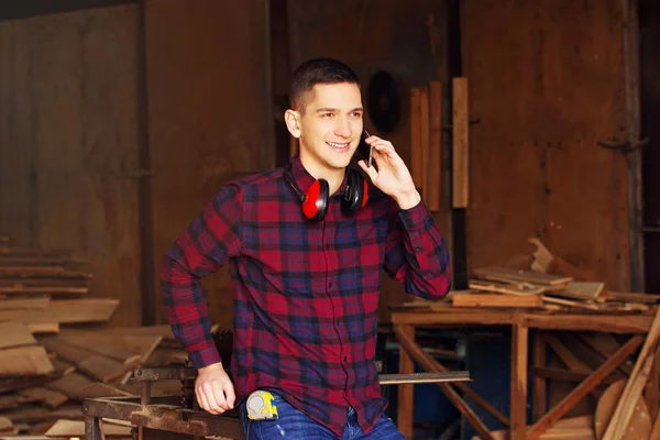 Smiling workman dressed in the checkered shirt talking the phone at the sawmill. Timbers on background.