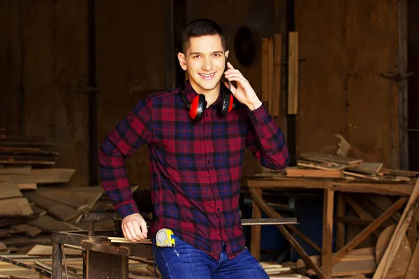 Smiling workman dressed in the checkered shirt talking the phone at the sawmill. Timbers on background.