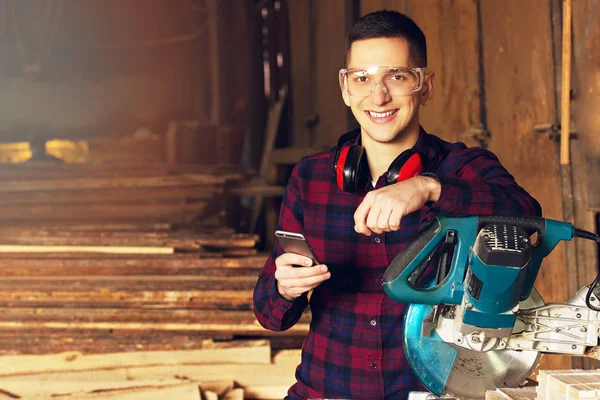 Smiling workman dressed in the checkered shirt and wearing protective glasses using his phone near circular saw. Timbers on background.