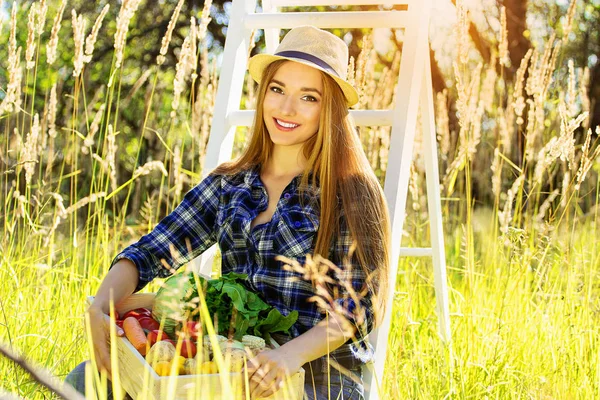 Menina sorridente bonita e jovem em chapéu mantendo caixa de madeira cheia de legumes. Colheita de verão . — Fotografia de Stock