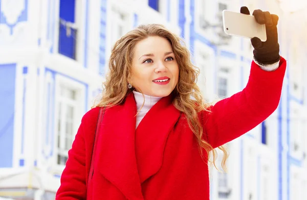 Fiatal, szép és stílusos szőke haj lány piros kabát, vesz egy selfie őt telefonon. Női divat. — Stock Fotó