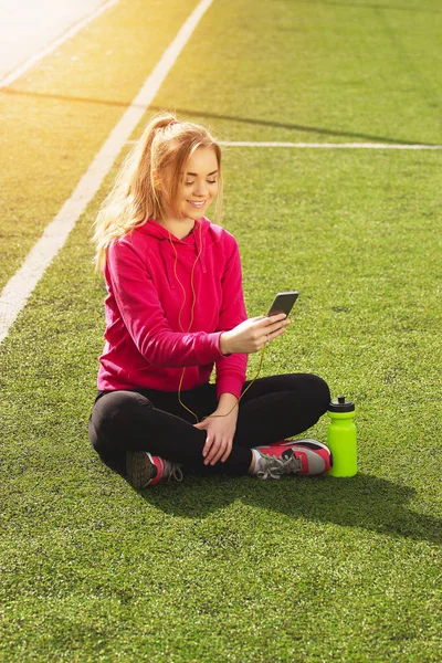 Fiatal gyönyörű szőke haja fitness lány használ neki telefont, a stadion. Nyári sport tevékenység. — Stock Fotó