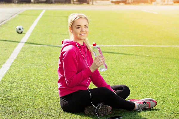 Szomjas fiatal gyönyörű szőke haja fitness lány ivóvíz a stadion. Nyári sport tevékenység. — Stock Fotó