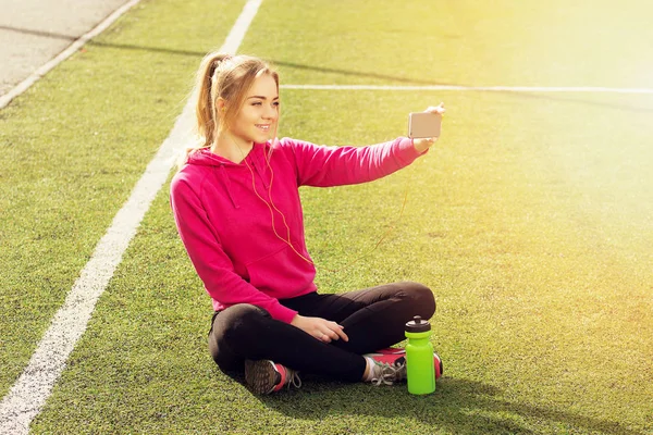 Fiatal gyönyörű szőke haja fitness lány csinál vele telefonon, a stadion selfie. Nyári sport tevékenység. — Stock Fotó