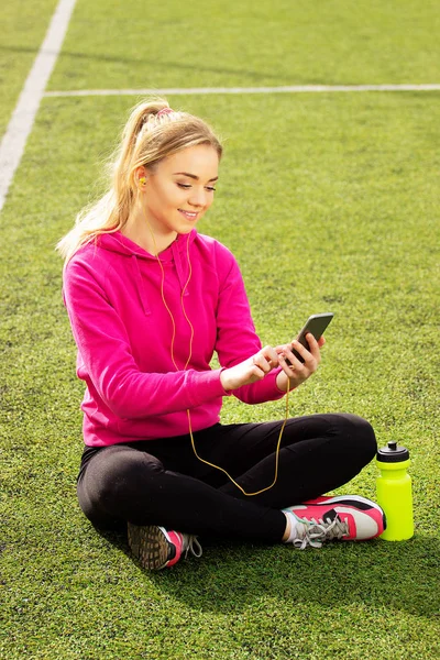 Fiatal gyönyörű szőke haja fitness lány használ neki telefont, a stadion. Nyári sport tevékenység. — Stock Fotó