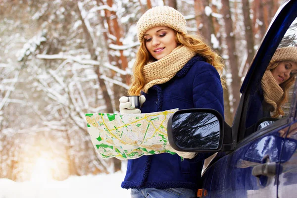 Beautiful young smiling girl in blue jacket standing near a car, holding map in hand and drinking tea. Travel girl.