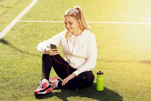 Fiatal gyönyörű szőke haja fitness lány használ neki telefont, a stadion. Nyári sport tevékenység. Zöld stadion fű háttér. — Stock Fotó