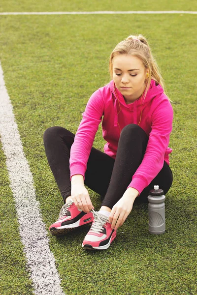 Fiatal gyönyörű szőke haj fitness lány kapcsolatok cipőfűző a stadion. Nyári sport tevékenység. Zöld stadion fű háttér. — Stock Fotó