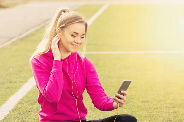 Portré, fiatal, gyönyörű szőke haja fitness lány használ neki telefont, a stadion. Nyári sport tevékenység. Zöld stadion fű háttér. — Stock Fotó