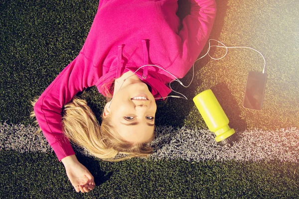 Porträt der jungen schönen blonden Haare Fitness-Mädchen liegt auf grünem Stadionrasen mit Telefon und Sportflasche in ihrer Nähe. Sommersport. Grüner Stadionrasen auf Hintergrund. — Stockfoto