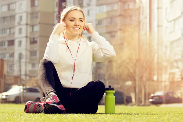 Junge schöne blonde Haare Fitness-Mädchen sitzt auf grünem Stadionrasen und hört Musik. Sommersport. — Stockfoto