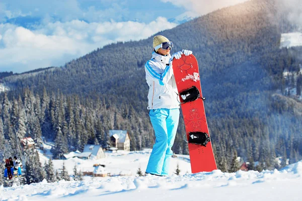 Krásná mladá dívka v bílém plášti, modré lyžařské kalhoty a brýle na její hlavy stojící s snowboard v zasněžených horách. Zimní sporty. — Stock fotografie