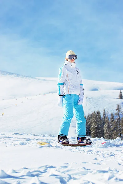 Junges schönes Mädchen in weißer Jacke, blauer Skihose und Brille auf dem Kopf, das auf dem Snowboard in den verschneiten Bergen reitet. Wintersport. — Stockfoto