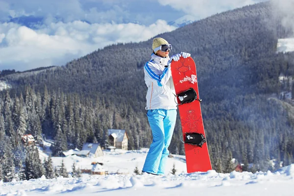 Krásná mladá dívka v bílém plášti, modré lyžařské kalhoty a brýle na její hlavy stojící s snowboard v zasněžených horách. Zimní sporty. — Stock fotografie