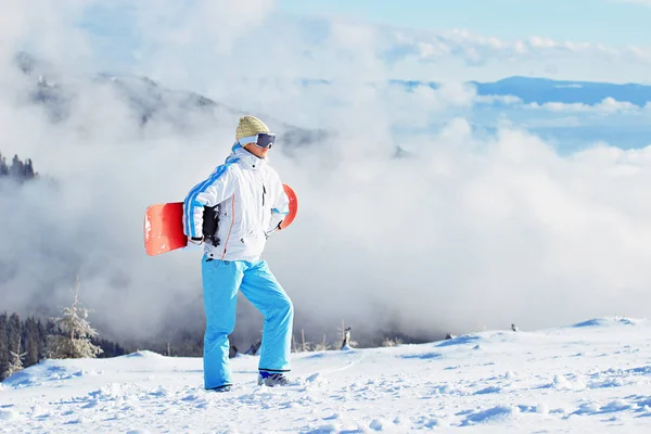 Όμορφη κοπέλα με άσπρο σακάκι, μπλε παντελόνι του σκι και τα googles στο κεφάλι στέκεται με snowboard στα χιονισμένα βουνά. Χειμερινά σπορ. — Φωτογραφία Αρχείου