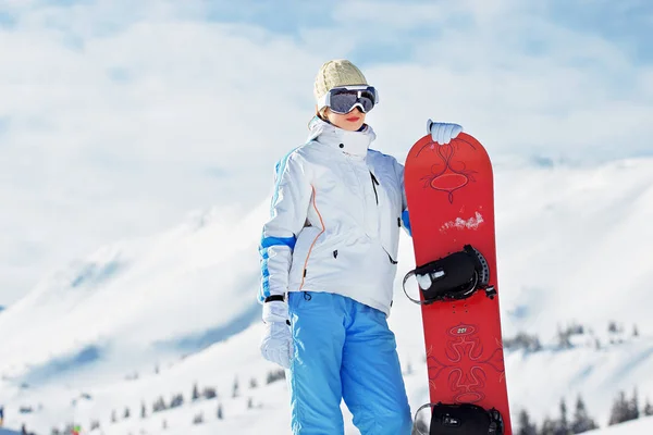 Ung vacker flicka i vit jacka, blå skidbyxor och googles på sitt huvud står med snowboard i de snöklädda bergen. Vintersport. — Stockfoto