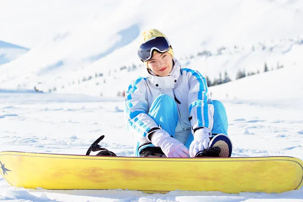 Krásná mladá dívka v bílém plášti, modré lyžařské kalhoty a brýle na hlavě sedí s snowboard v zasněžených horách. Zimní sporty. — Stock fotografie