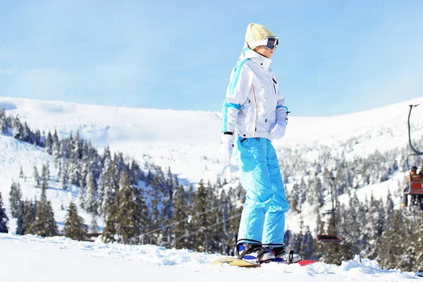 Krásná mladá dívka v bílém plášti, modré lyžařské kalhoty a brýle na hlavě, jízda na snowboardu v zasněžených horách. Zimní sporty. — Stock fotografie