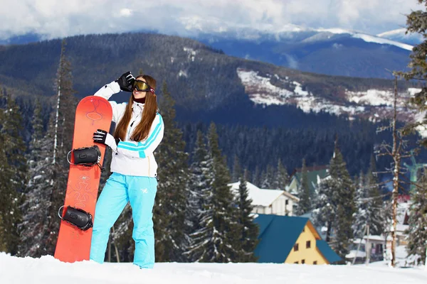 Krásná mladá usměvavá dívka v bílém plášti, modré lyžařské kalhoty a brýle na její hlavy stojící s snowboard v zasněžených horách. Zimní sporty. — Stock fotografie