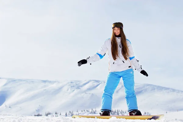 Jonge mooi lachende meisje in een witte jas, blue ski broeken en googles op haar hoofd rijden op snowboard in de besneeuwde bergen. Wintersport. — Stockfoto