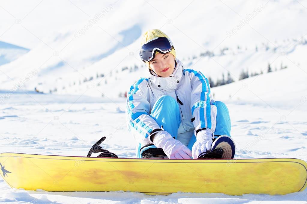 Young beautiful girl in white jacket, blue ski pants and googles on her head sitting with snowboard in the snowy mountains. Winter sports.