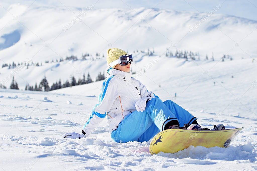 Young beautiful girl in white jacket, blue ski pants and googles on her head sitting with snowboard in the snowy mountains. Winter sports.