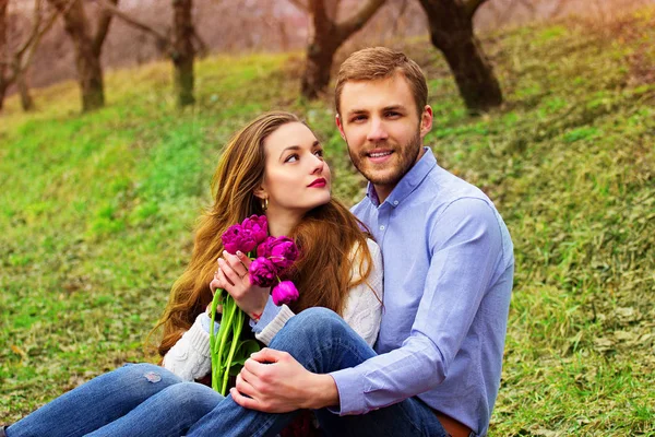 Casual happy νεαρό ζευγάρι. Πορτρέτο του hipster γενειοφόρος άνδρας κάθεται μαζί με την όμορφη κοπέλα με λουλούδια στον κήπο. — Φωτογραφία Αρχείου
