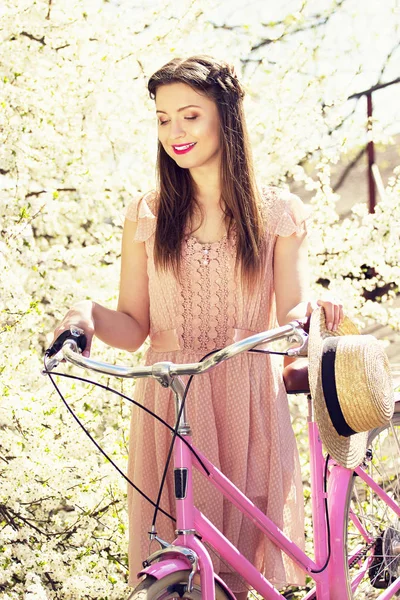 春の美しさ。美しい長い髪の少女の肖像画を着たレトロな自転車とピンクのドレスの立っています。背景の花. — ストック写真