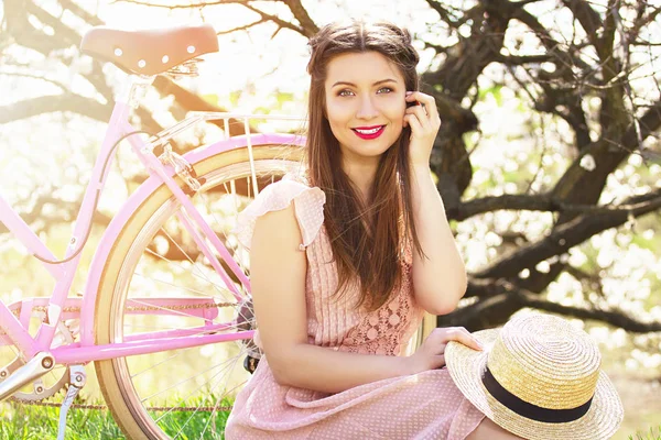 Retro style. Portrait of smiling young and beautiful long-haired girl in pink dress sitting with retro bike on flower background. — Stock Photo, Image