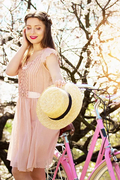Humor de primavera. Retrato de sorrir menina bonita e jovem de cabelos compridos em vestido rosa de pé com bicicleta retro. Fundo flor . — Fotografia de Stock
