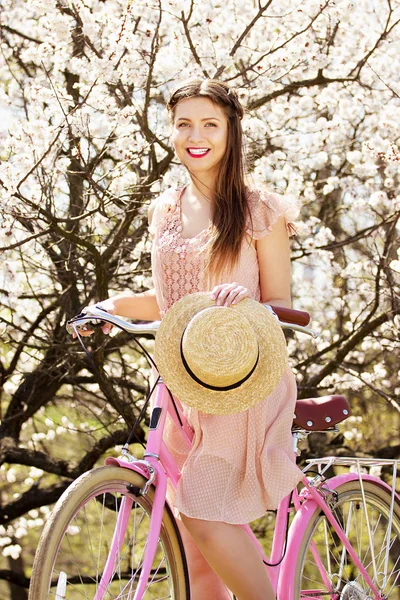 Montar en bicicleta. Retrato de una joven sonriente y hermosa chica de pelo largo en vestido rosa montando una bicicleta retro y sosteniendo el sombrero sobre fondo de flores . — Foto de Stock