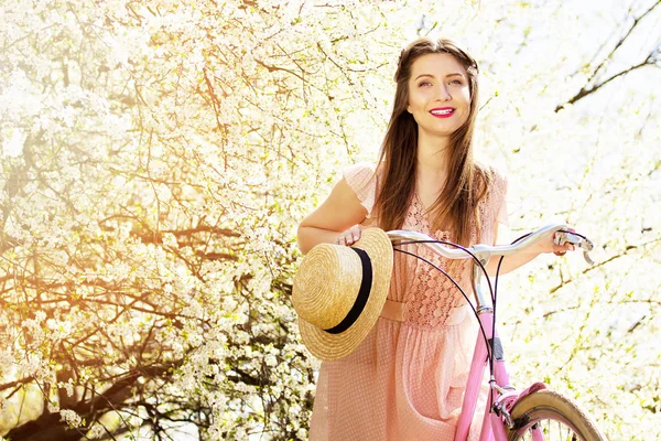 Flores à tua volta. Retrato de sorrir jovem e bela menina de cabelos compridos em vestido rosa de pé com bicicleta retro no fundo da flor . — Fotografia de Stock